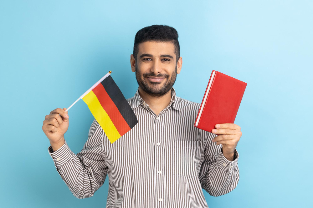 portrait-positive-smiling-man-showing-holding-book-german-flag-dreaming-study-germany-education-abroad-wearing-striped-shirt-indoor-studio-shot-isolated-blue-background